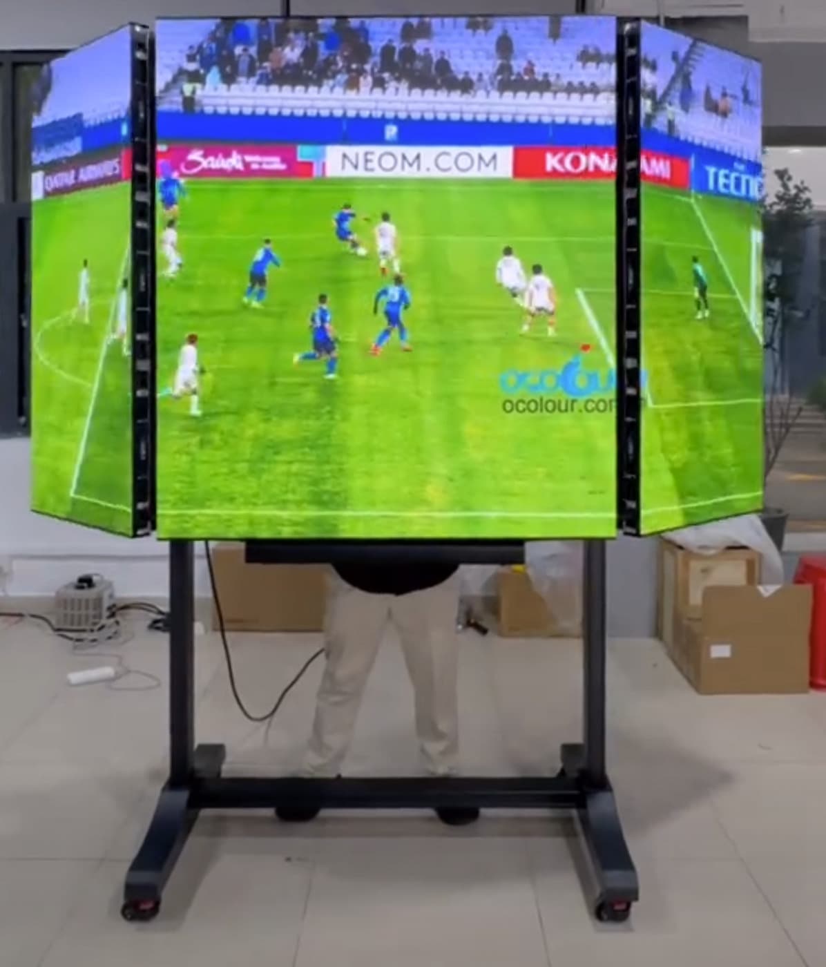 Mobiler LED-TV mit zwei teilweise eingeklappten Seitenflächen, Fußballbild und schwarzem Rollständer. Standbild aus einem Video.