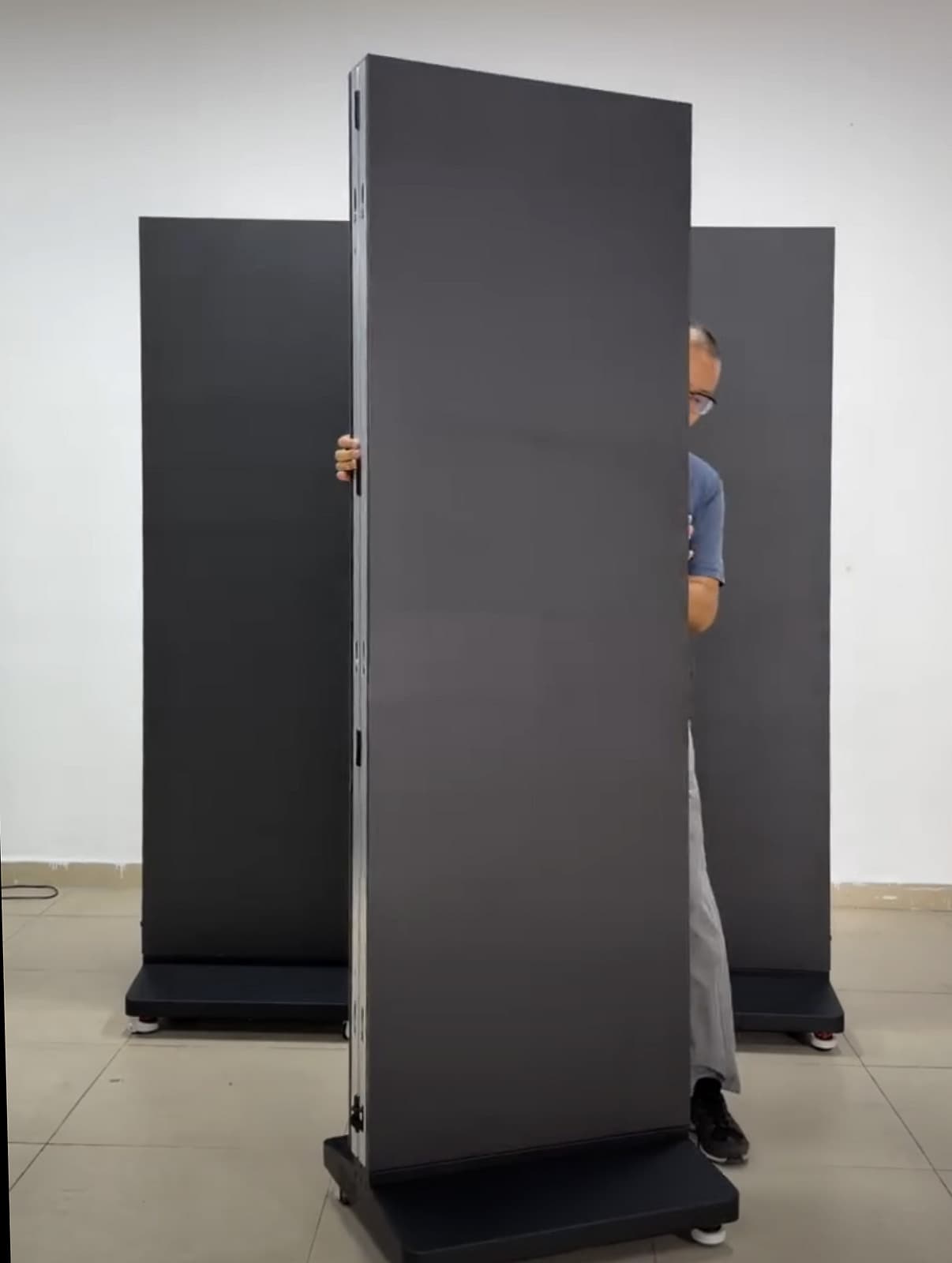 Vorderseite eines schmalen LED-Aufstellers mit dunkler Bildfläche, seitlicher Gehäusekante und Rollfuß vor zwei weiteren Geräten. Standbild aus einem Video.