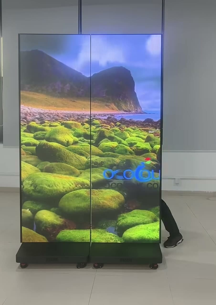 Zwei gekoppelte LED-Faltposter mit Landschaftsmotiv vor einer Hallenwand und sichtbaren Beinen rechts