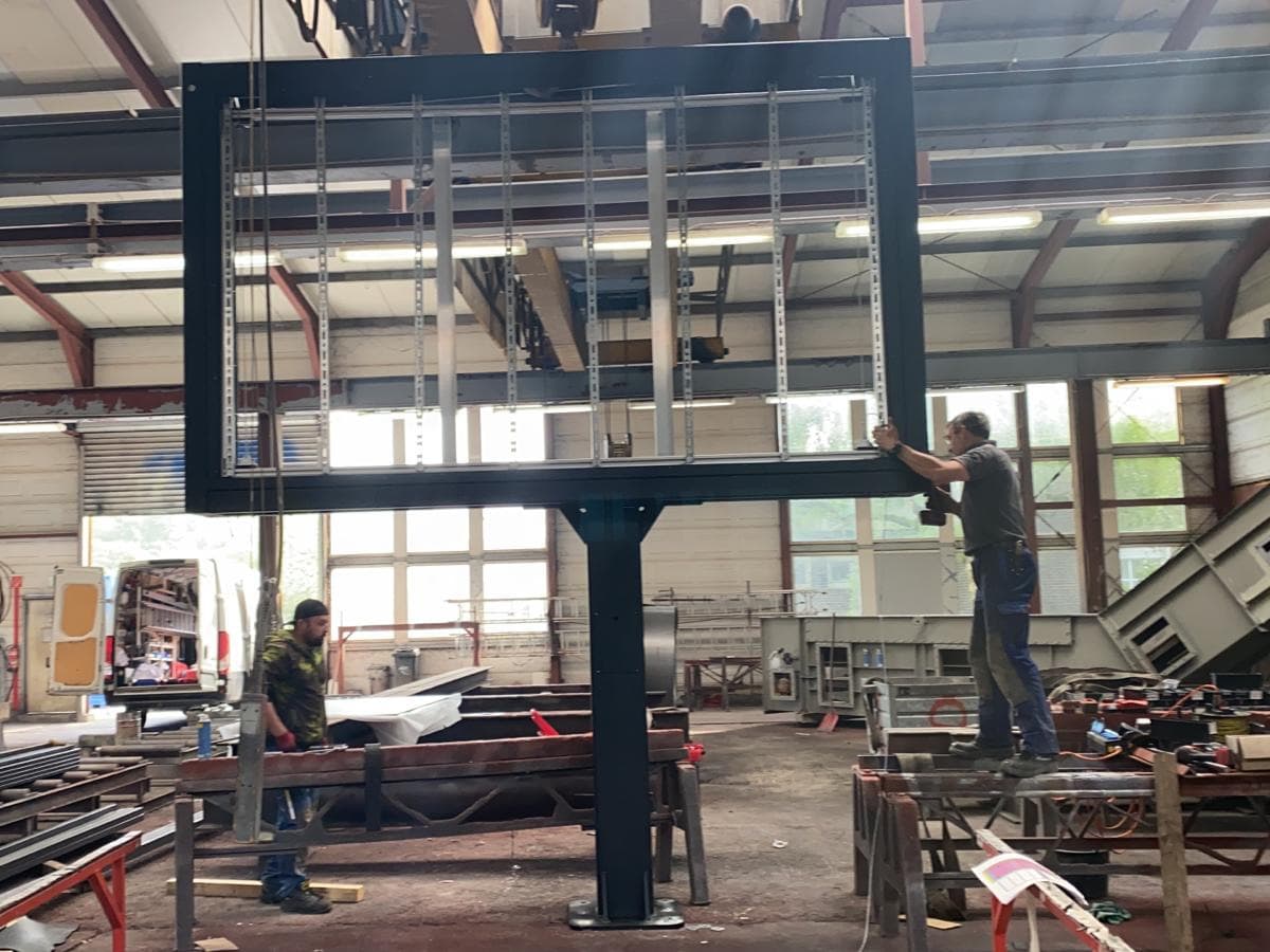Stahlrahmen auf einem Einzelmast bei der Montage durch zwei Personen in einer Metallbauhalle